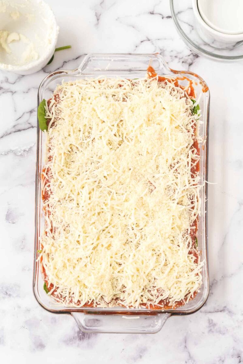 A glass baking dish filled with unbaked lasagna with spinach and ricotta, topped with a generous layer of shredded cheese, sits on a white marble surface next to empty bowls.