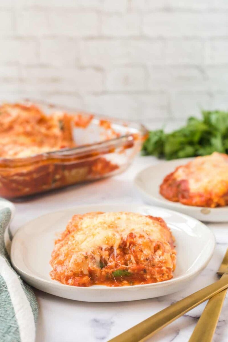A slice of cheesy baked lasagna with spinach and ricotta is served on a white plate, with another plate and a glass baking dish of lasagna in the background. Gold-colored cutlery and a green towel are placed nearby.