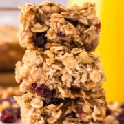 Three oat breakfast bars with visible cranberries are stacked on top of each other, surrounded by scattered nuts and berries, with a blurred background featuring a yellow container.