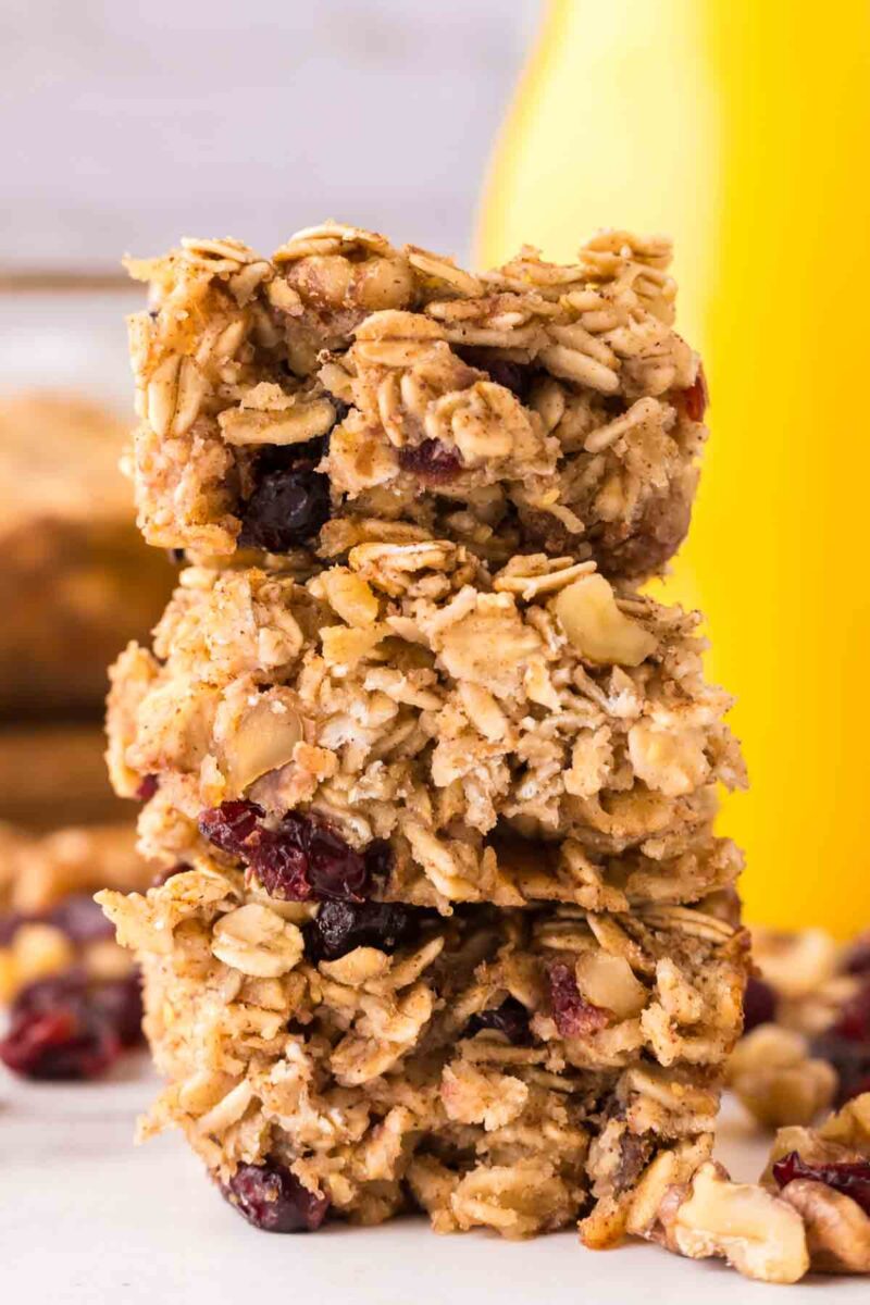 Three oat breakfast bars with visible cranberries are stacked on top of each other, surrounded by scattered nuts and berries, with a blurred background featuring a yellow container.