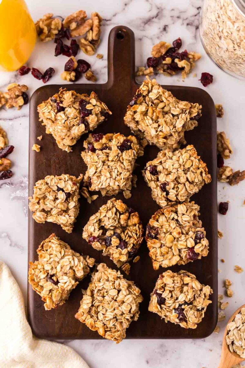 A dark wooden board holds several oat breakfast bars with cranberries, surrounded by scattered oats, walnuts, and dried cranberries on a white marble surface. A jar of oats and a yellow cup are visible nearby.
