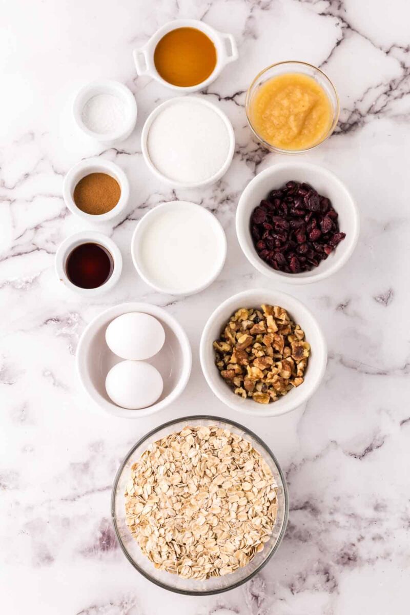 Various baking ingredients for oat breakfast bars are arranged on a white marble surface, including rolled oats, eggs, walnuts, dried cranberries, milk, applesauce, vanilla, cinnamon, sugar, baking powder, and salt in small bowls.