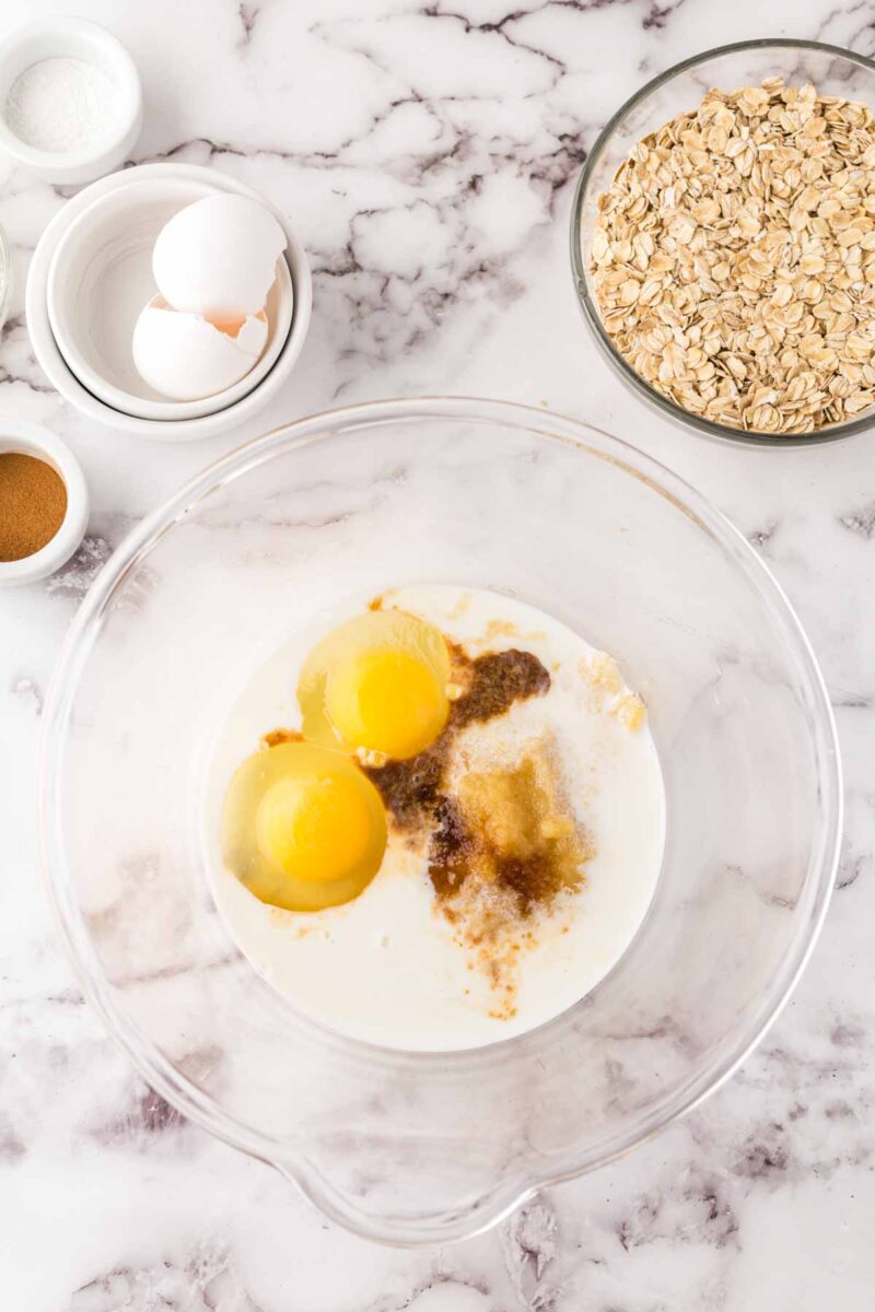 A clear mixing bowl with eggs, milk, brown sugar, and vanilla sits on a marble countertop, surrounded by bowls of oats, ground cinnamon, salt, and whole eggs—perfect ingredients for whipping up homemade oat breakfast bars.