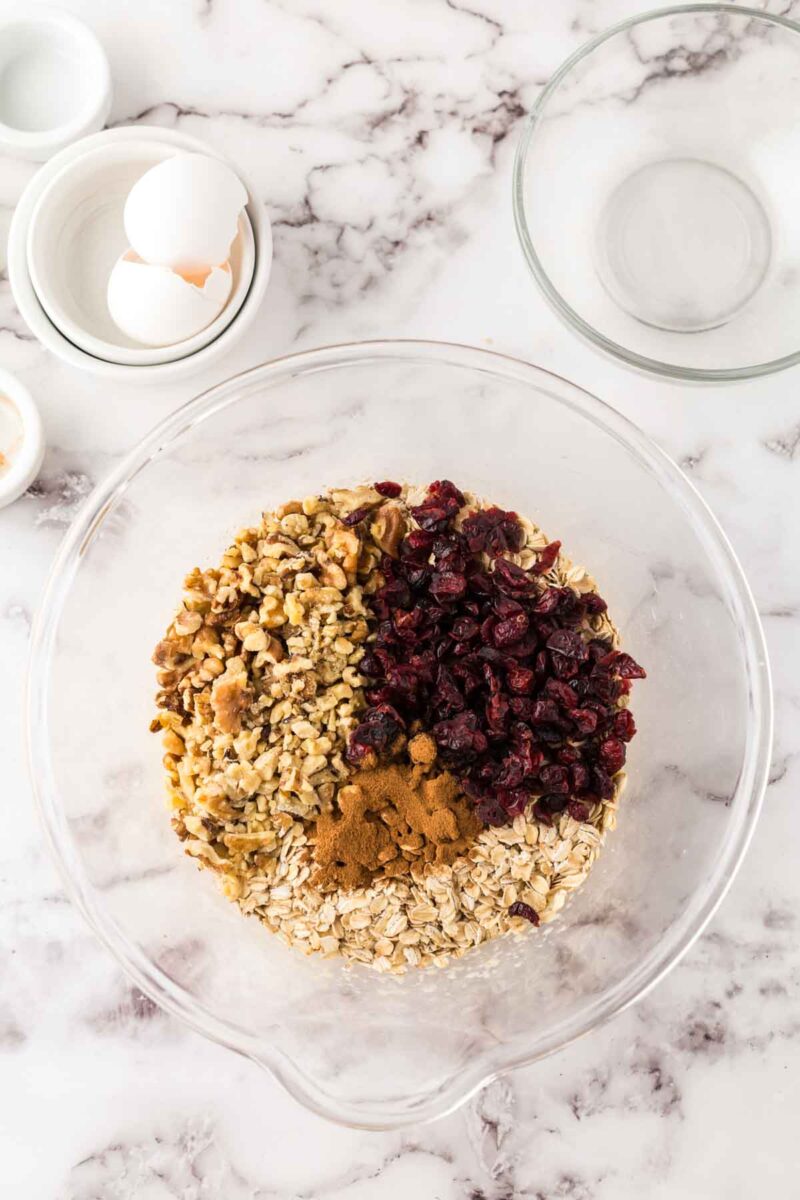 A glass bowl containing oats, chopped walnuts, dried cranberries, and a pile of cinnamon sits on a marble countertop next to empty bowls and cracked eggshells—perfect ingredients for homemade oat breakfast bars.