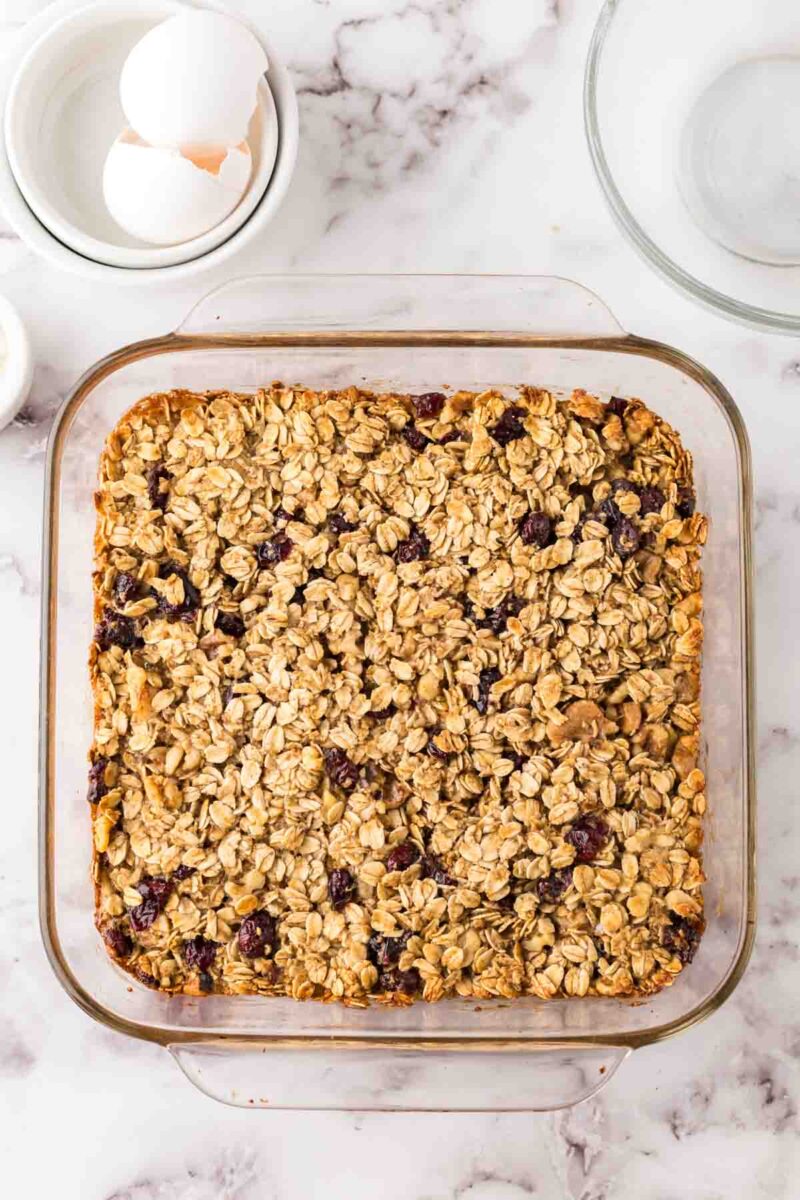 A square glass baking dish filled with oat breakfast bars topped with oats and berries sits on a marble countertop. Nearby are a bowl with cracked eggs and an empty glass mixing bowl.
