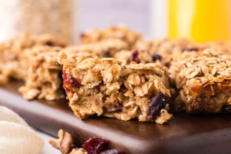 Close-up of homemade oat breakfast bars with visible oats and dried cranberries, arranged on a wooden board with a blurred background.
