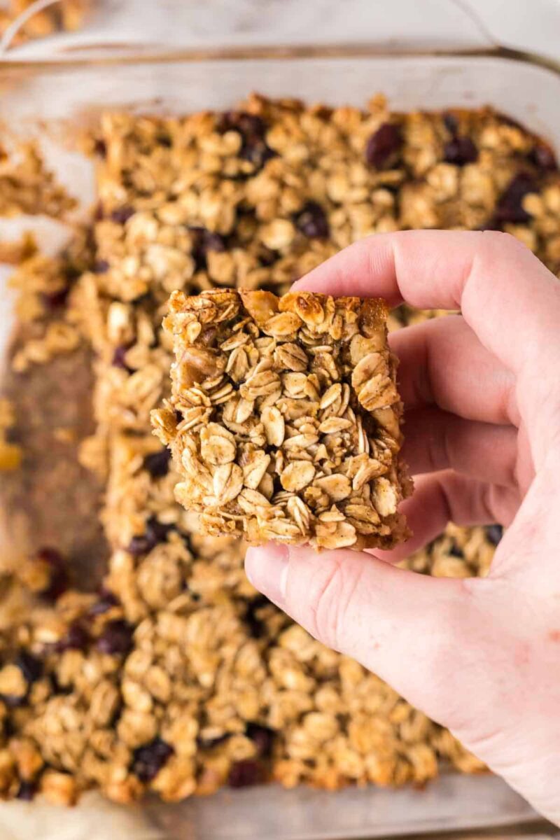 A hand holding a square oat breakfast bar with visible oats and raisins, with more oat bars in a glass baking dish in the background.