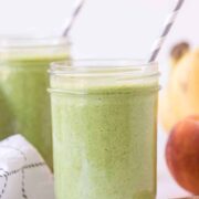 A mason jar filled with a simple green smoothie recipe and a striped straw sits on a white surface. In the background, there’s another jar, a peach, a banana, and a cloth napkin.