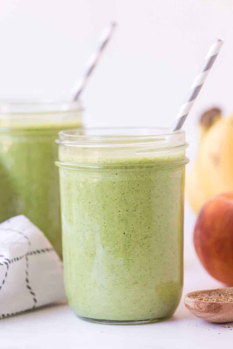 A mason jar filled with a simple green smoothie recipe and a striped straw sits on a white surface. In the background, there’s another jar, a peach, a banana, and a cloth napkin.