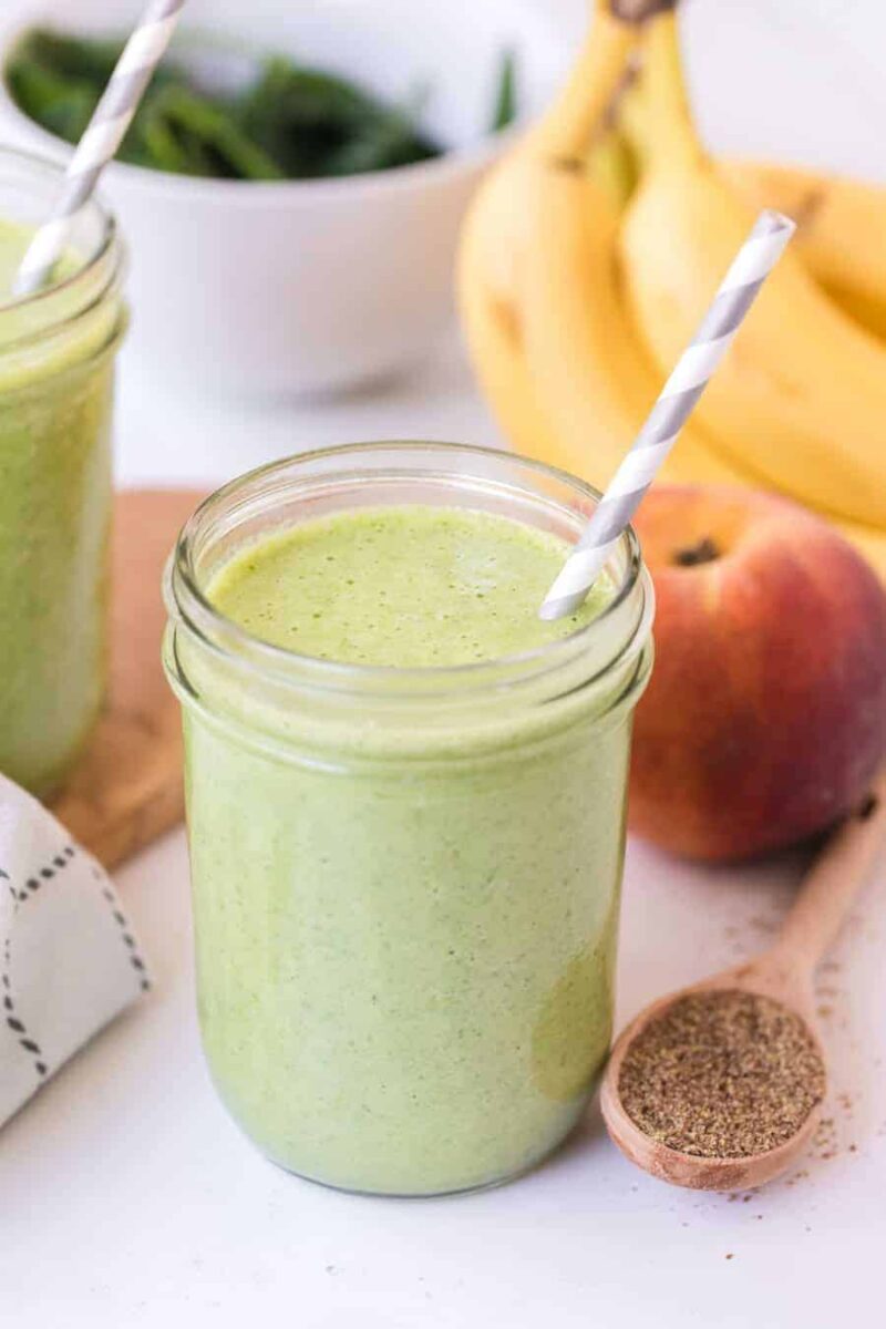 A mason jar filled with a simple green smoothie recipe and a striped paper straw, with bananas, a peach, a bowl of spinach, and a spoonful of ground flaxseed in the background.
