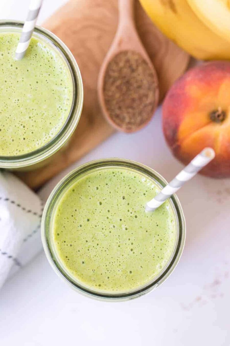 Two glasses of green smoothie with striped straws sit beside a peach, a banana, a wooden spoon filled with ground flaxseed, and a white towel—perfect for anyone seeking a simple green smoothie recipe.