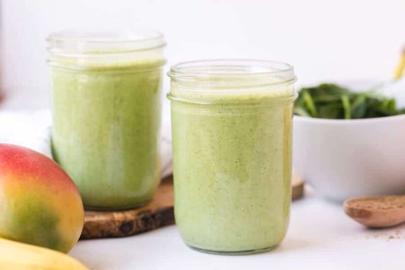 Two mason jars filled with a vibrant green smoothie sit on a light surface. Nearby, a mango, banana, leafy greens, and a spoon hint at the simple green smoothie recipe behind this refreshing drink.
