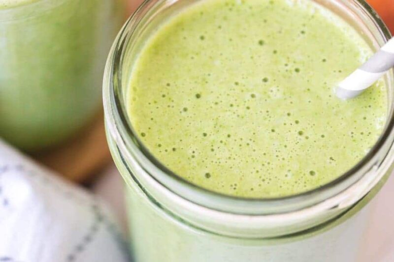 A close-up of a glass filled with a creamy green smoothie made from a simple green smoothie recipe, with a white-striped straw inserted. The surface is frothy, and another glass and a cloth are partially visible in the background.
