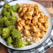 A plate of brown rice topped with teriyaki chicken pieces, served with steamed broccoli, sits on a white plate with a black rim. The dish showcases a delicious teriyaki chicken recipe, artfully arranged on a checkered napkin over a wooden surface.