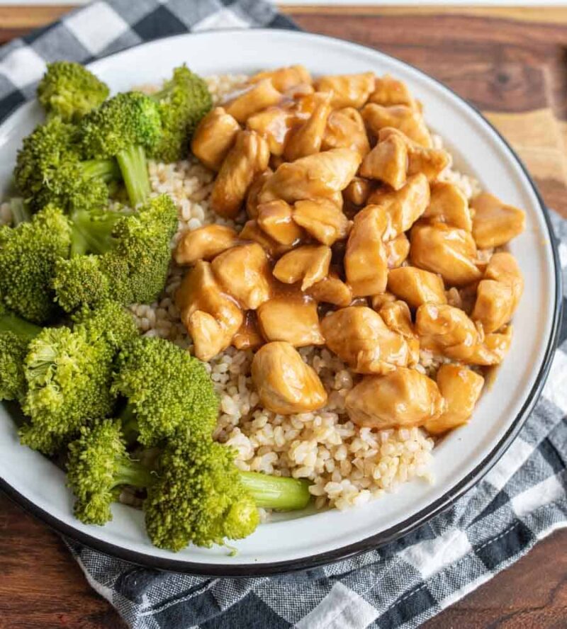 A plate of brown rice topped with teriyaki chicken pieces, served with steamed broccoli, sits on a white plate with a black rim. The dish showcases a delicious teriyaki chicken recipe, artfully arranged on a checkered napkin over a wooden surface.