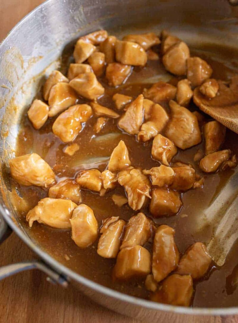 Chunks of chicken coated in a glossy brown sauce are being stirred in a metal pan with a wooden spoon, creating an appetizing teriyaki chicken recipe on a wooden surface.