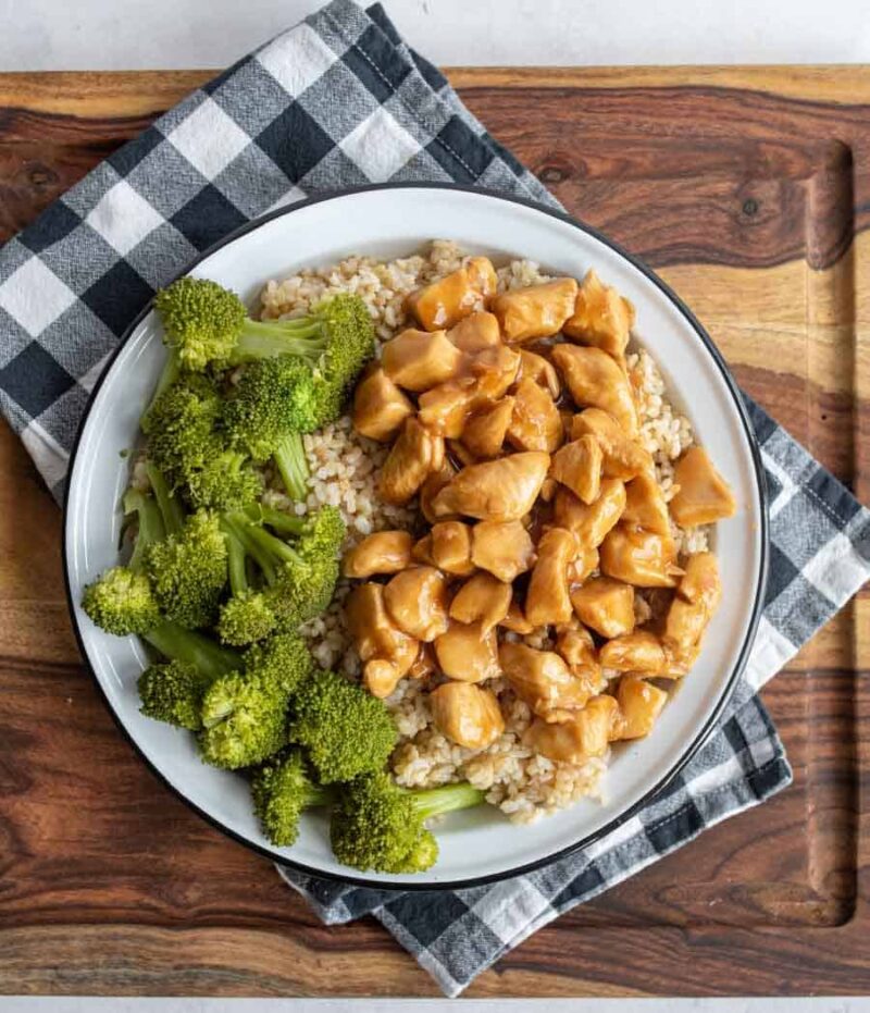 A plate of brown rice topped with flavorful teriyaki chicken pieces and steamed broccoli, served on a black and white checkered napkin atop a wooden cutting board—a perfect meal for trying out your favorite teriyaki chicken recipe.