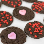 Decorated Valentine Day cookies: Chocolate cookies topped with pink and yellow heart-shaped candies and small red heart-shaped sprinkles, arranged on a white surface.