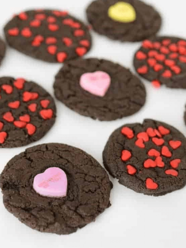 Decorated Valentine Day cookies: Chocolate cookies topped with pink and yellow heart-shaped candies and small red heart-shaped sprinkles, arranged on a white surface.