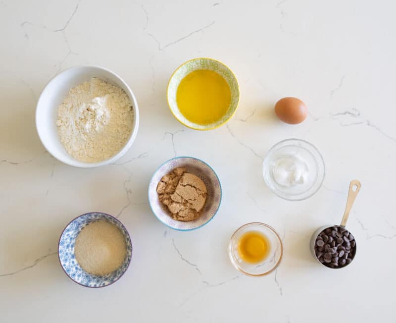 Several small bowls and a measuring cup with baking ingredients—flour, melted butter, an egg, brown sugar, white sugar, granulated sugar, vanilla extract, and chocolate chips—are arranged on a white marble surface.