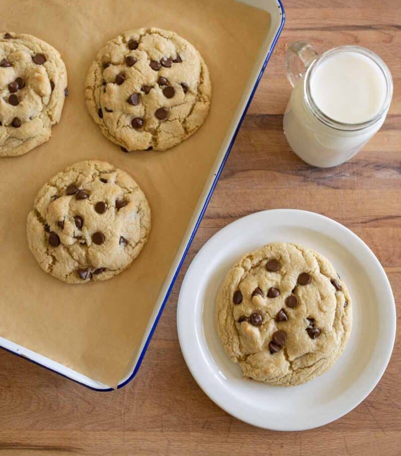 A plate with a large chocolate chip cookie sits next to a tray of three more cookies on parchment paper. A mason jar glass of milk is nearby on a wooden table.