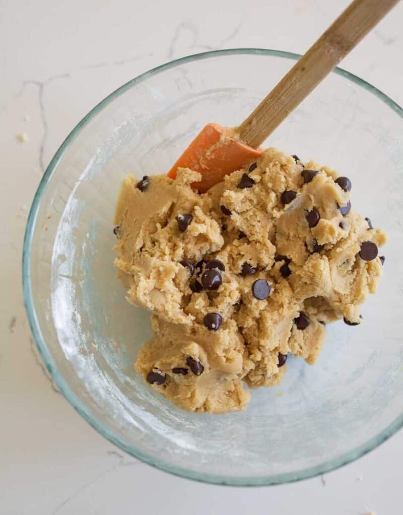 A glass bowl filled with chocolate chip cookie dough, mixed with a wooden-handled orange spatula, sits on a white countertop.