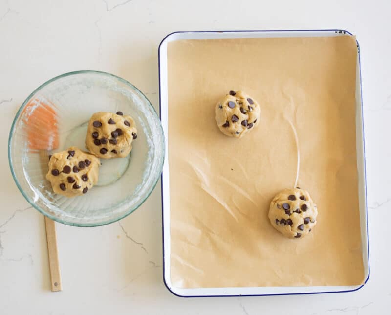 A glass bowl with cookie dough and a spatula sits next to a baking tray lined with parchment paper, holding three dough balls with visible chocolate chips, ready to be baked.