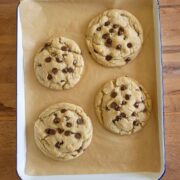 Four large chocolate chip cookies on a sheet of parchment paper in a rectangular baking pan, resting on a wooden surface.