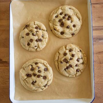 Four large chocolate chip cookies on a sheet of parchment paper in a rectangular baking pan, resting on a wooden surface.
