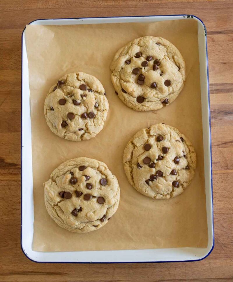 Four large chocolate chip cookies on a sheet of parchment paper in a rectangular baking pan, resting on a wooden surface.