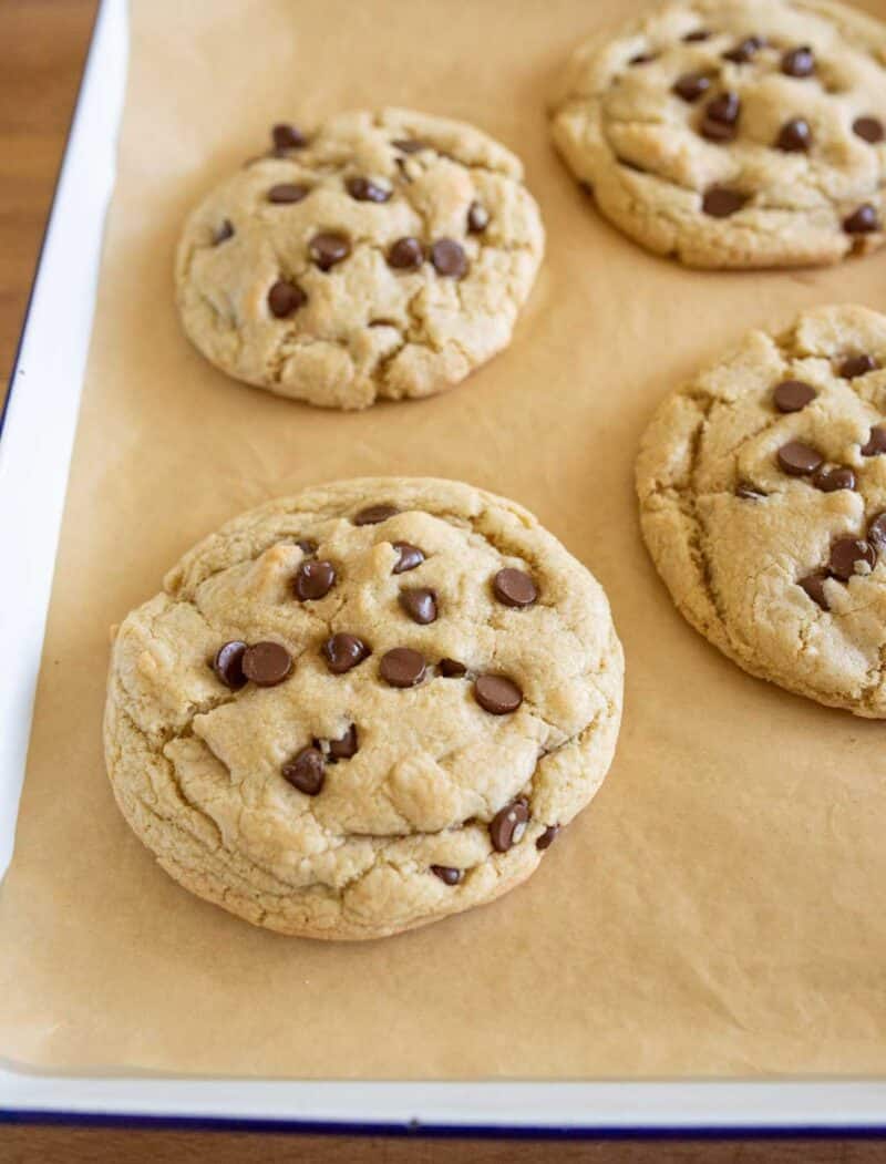 Four large chocolate chip cookies rest on a sheet of parchment paper in a baking tray. The cookies are golden brown with melty chocolate chips scattered on top.