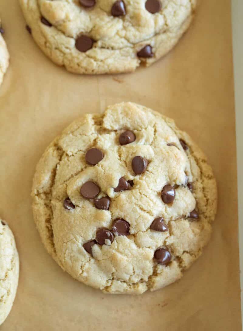 A large, soft chocolate chip cookie with scattered chocolate chips sits on a sheet of brown parchment paper.