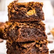 Three thick, fudgy chocolate brownies with a swirl of peanut butter are stacked on top of each other on a piece of parchment paper, with a blurred background.