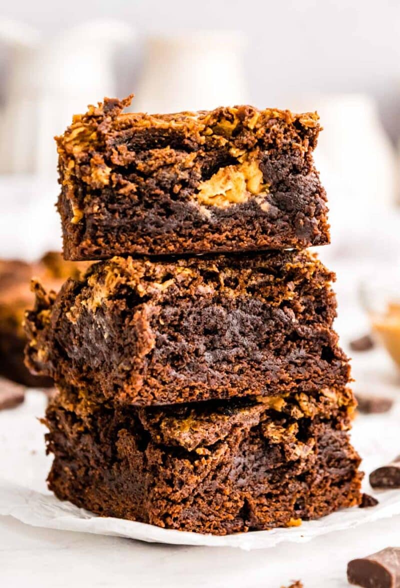 Three thick, fudgy chocolate brownies with a swirl of peanut butter are stacked on top of each other on a piece of parchment paper, with a blurred background.