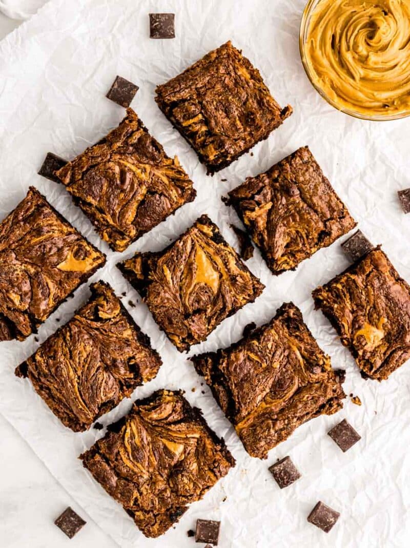 Nine brownies with a marbled swirl pattern, likely of chocolate and peanut butter, are arranged on crumpled parchment paper. Chocolate chunks and a bowl of creamy peanut butter are also visible.
