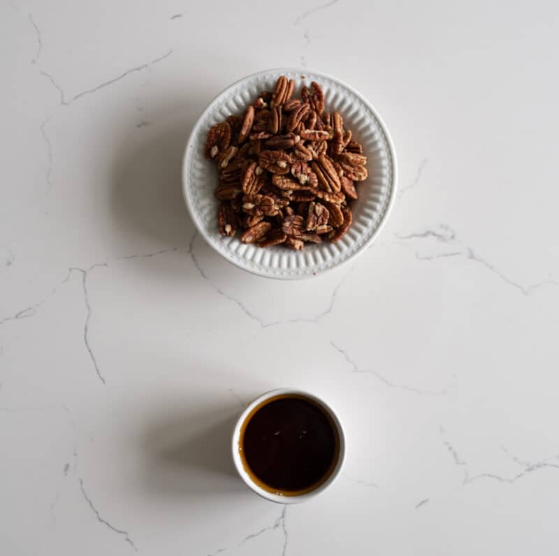 A white bowl filled with pecan halves and a small cup of dark liquid, possibly coffee or syrup, on a white, marble-patterned surface.