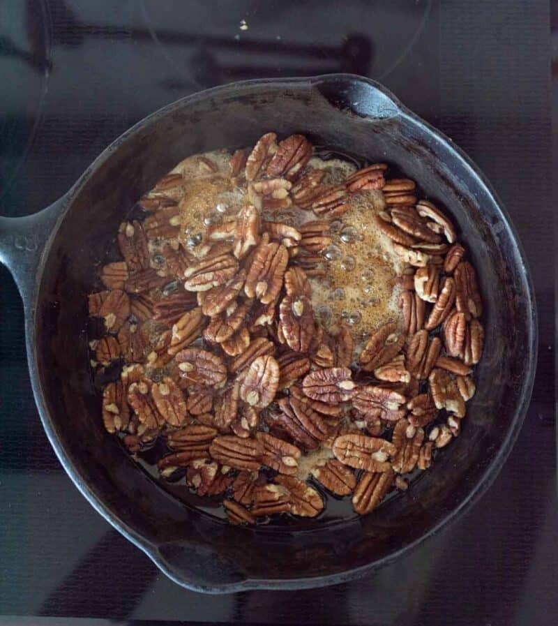 Pecan halves cooking in bubbling brown butter in a black cast iron skillet on a stovetop.
