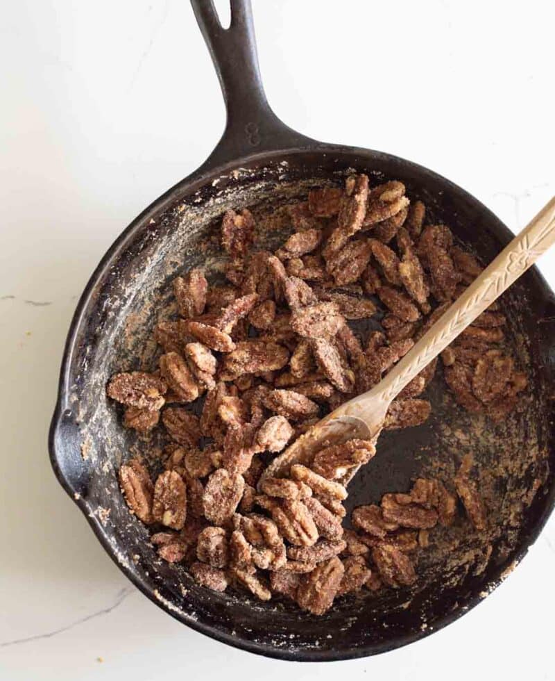 A cast iron skillet filled with sugared pecans being stirred with a wooden spoon, set on a white surface.