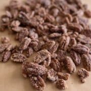 A close-up of candied pecans coated in sugar and spices, scattered on a sheet of brown parchment paper.