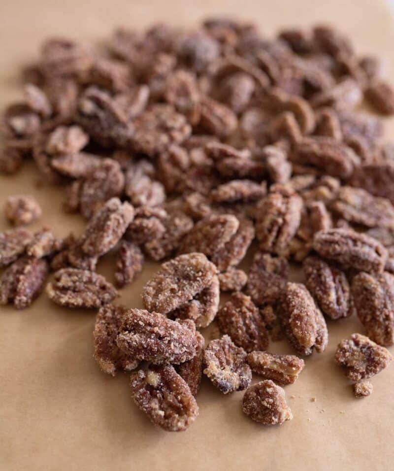 A close-up of candied pecans coated in sugar and spices, scattered on a sheet of brown parchment paper.