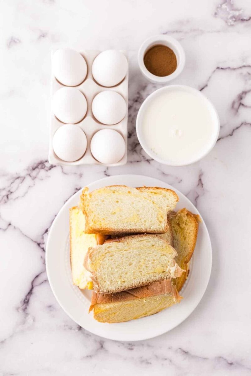 A plate with stacked slices of bread is on a marble surface, next to a carton holding six eggs, a small bowl of cinnamon, and a bowl of milk.