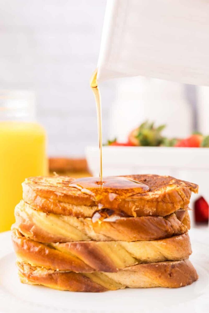 A stack of four slices of French toast on a white plate with syrup being poured on top. In the background, there is a glass of orange juice and a bowl of strawberries.