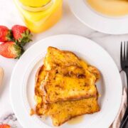 A plate with three slices of French toast drizzled with syrup, next to a fork and napkin. Fresh strawberries and a glass of orange juice are on the table, with another plate of French toast in the background.