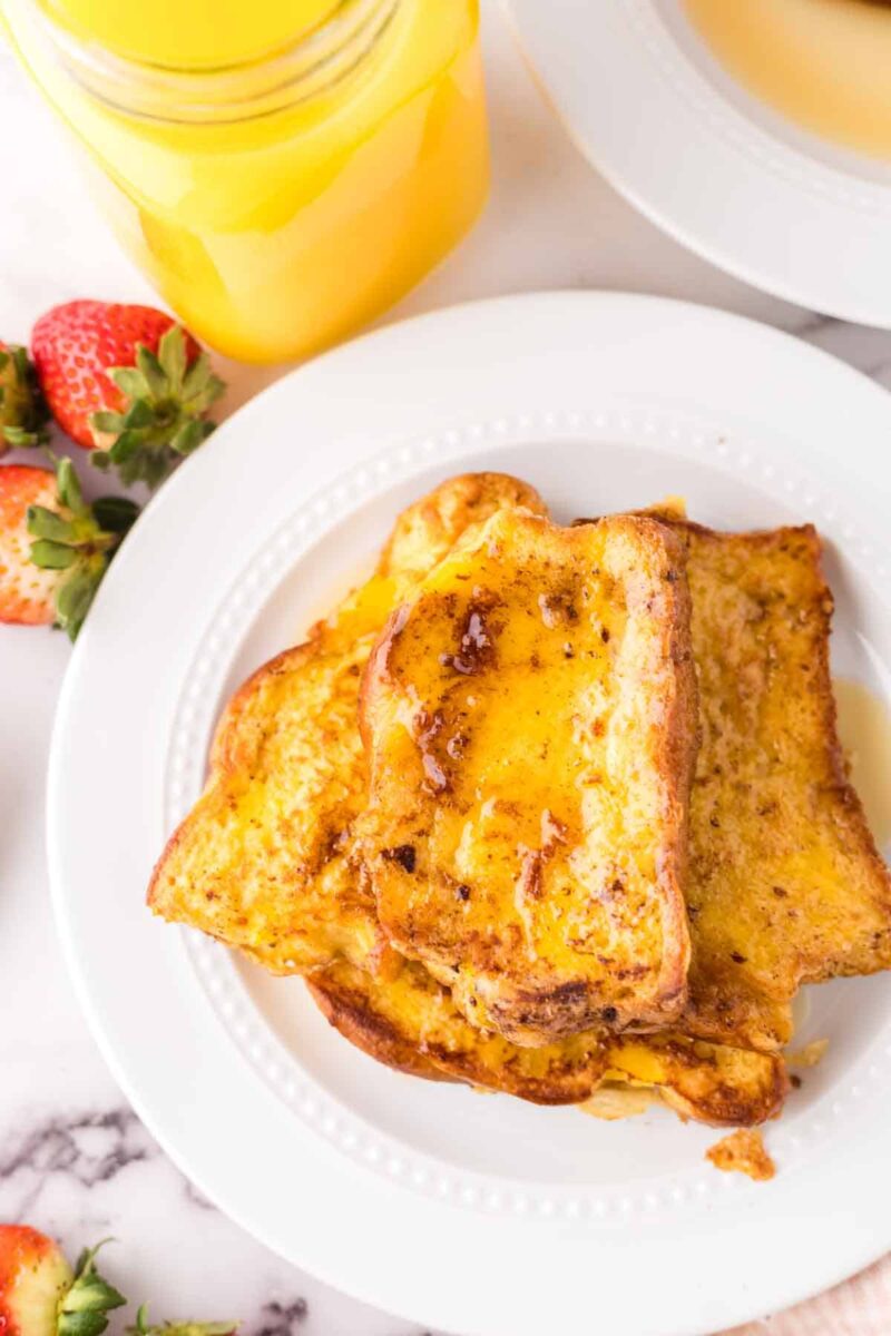 Three slices of French toast drizzled with syrup are served on a white plate. Fresh strawberries and a jar of orange juice are nearby on a marble surface.