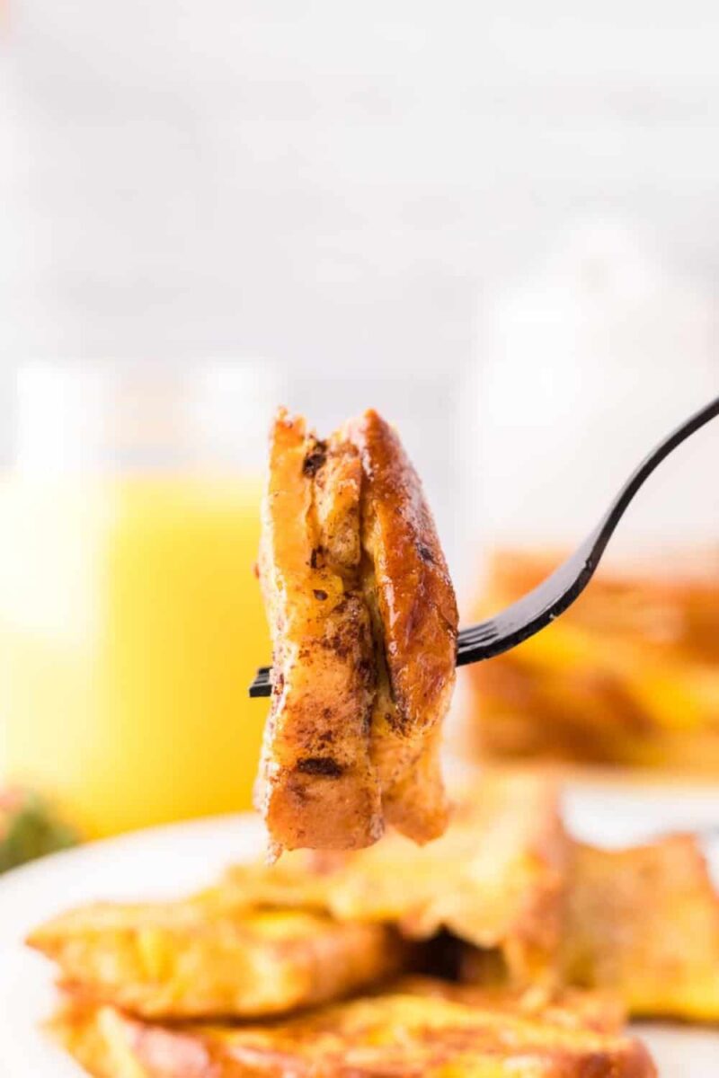 A close-up of a fork holding a bite-sized piece of stuffed French toast, with a blurred background showing more French toast and a glass of orange juice.