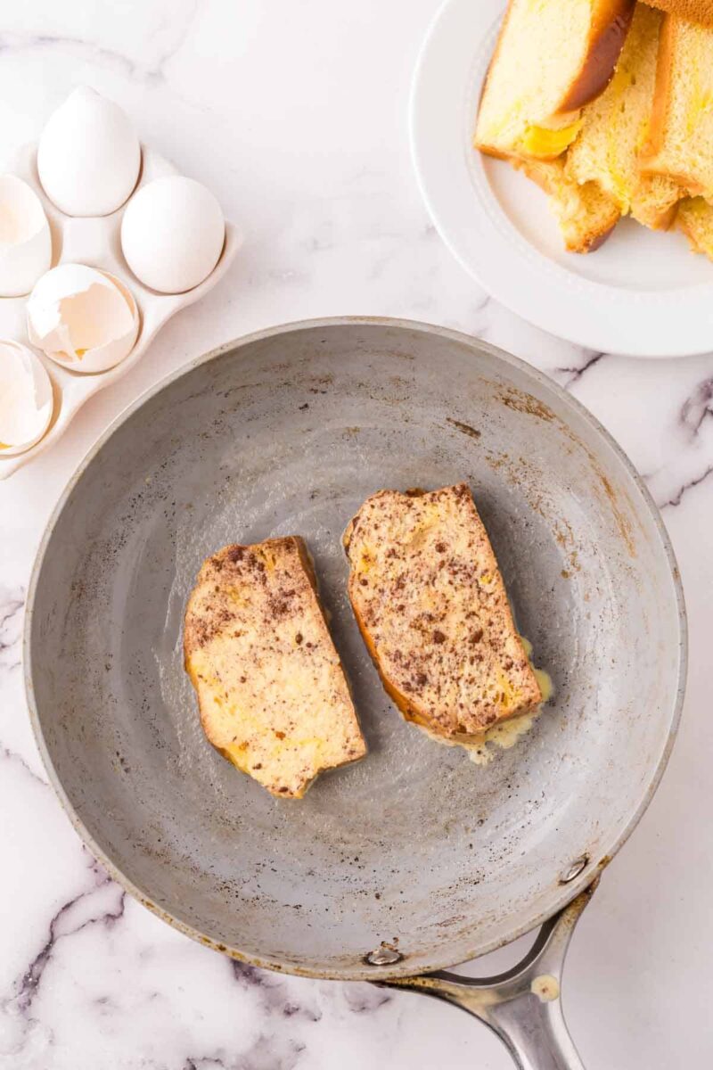 Two slices of French toast cooking in a metal frying pan, with a plate of sliced bread and cracked egg shells on a marble countertop nearby.