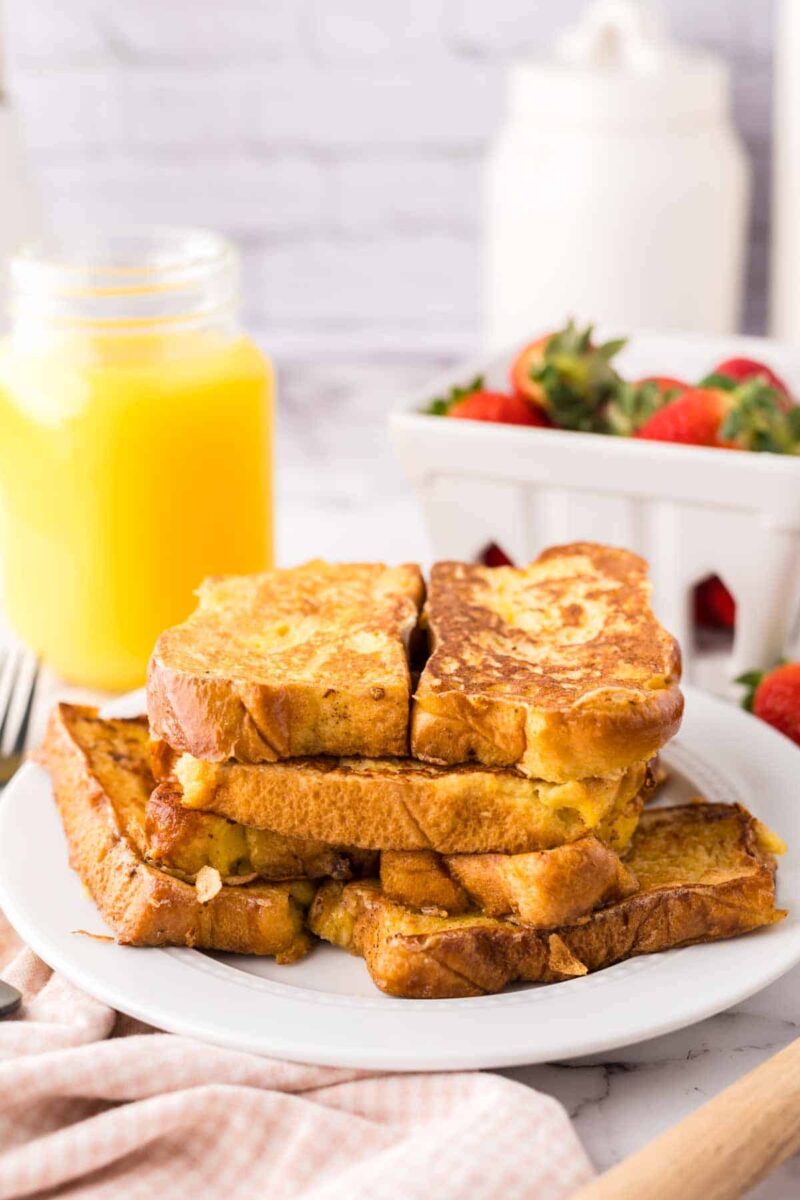 A plate of golden-brown French toast slices is stacked on a white plate. In the background, there is a jar of orange juice and a white basket filled with fresh strawberries. A fork and napkin are nearby.
