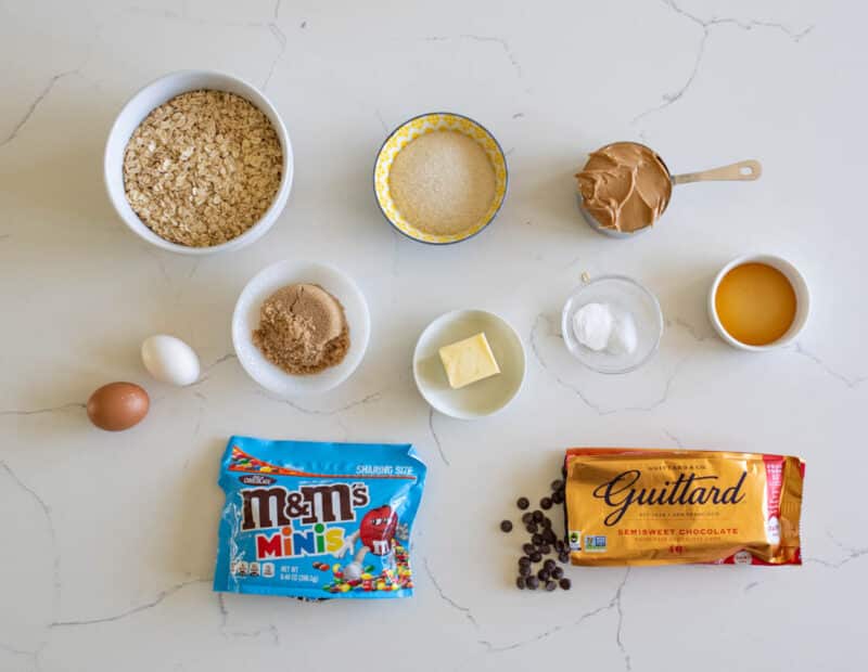 Ingredients for cookies arranged on a white countertop: oats, sugar, peanut butter, brown sugar, butter, vanilla, baking soda, salt, an egg, an egg white, a bag of mini M&M’s, a chocolate bar, and a few chocolate chips.
