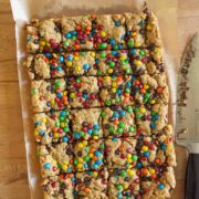 A tray of cookie bars topped with colorful candy-coated chocolates, cut into squares, sits on parchment paper on a wooden surface. A large knife with crumbs is placed next to the tray.