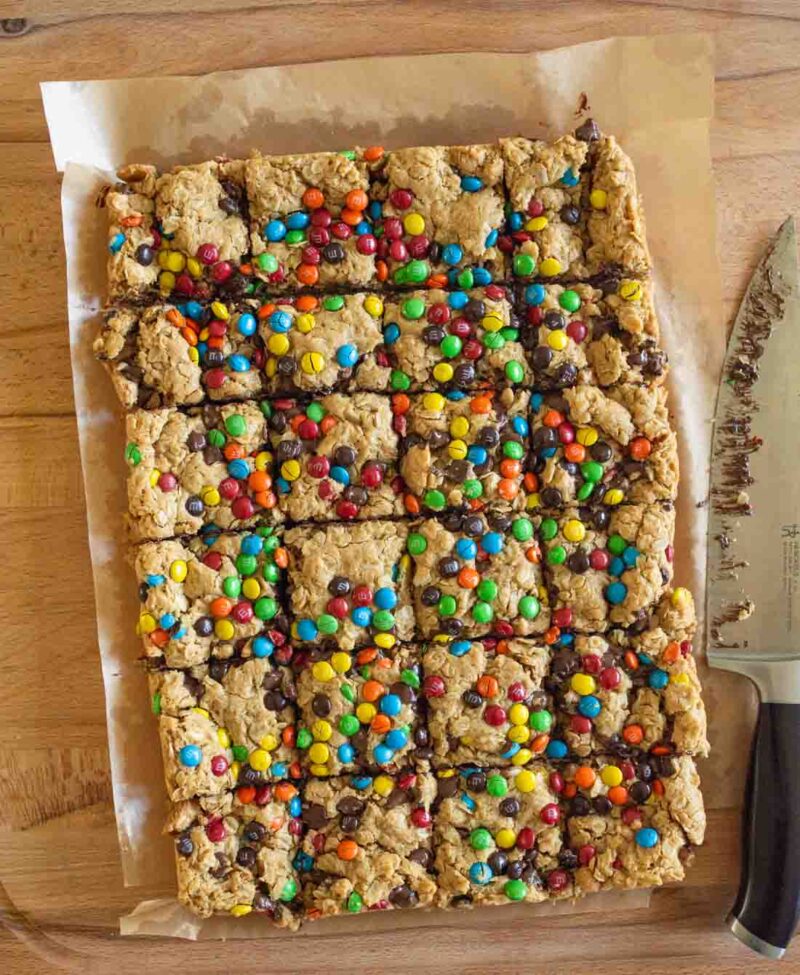A tray of cookie bars topped with colorful candy-coated chocolates, cut into squares, sits on parchment paper on a wooden surface. A large knife with crumbs is placed next to the tray.